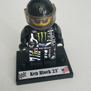 Può includere: Una minifigure Lego nera e verde del pilota di auto da corsa Ken Block, che indossa un casco nero con il logo Monster Energy e occhiali da sole. La minifigure è in piedi su una base nera con una bandiera a scacchi e il testo "Ken Block 23".