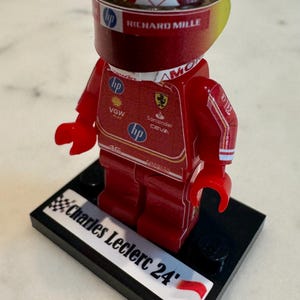 May include: A red and yellow Lego figurine of a race car driver wearing a red helmet with a white and yellow design. The figurine is standing on a black base with the words "Charles Leclerc 24" and a small red and white flag.