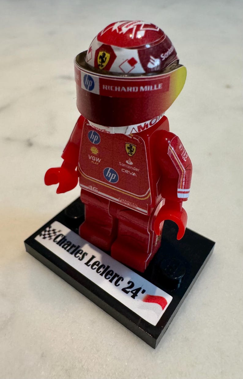 May include: A red and yellow Lego figurine of a race car driver wearing a red helmet with a white and yellow design. The figurine is standing on a black base with the words "Charles Leclerc 24" and a small red and white flag.