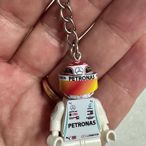 Könnte beinhalten: Schlüsselanhänger mit einer Lego-ähnlichen Rennfahrerfigur. Die weiße Figur trägt einen Helm mit dem "PETRONAS"-Logo, roten, gelben und weißen Details und der Nummer "44". Der Schlüsselanhänger ist an einer silbernen Kette befestigt.