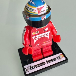 Può includere: Una statuetta Lego di un pilota di Formula 1, con una tuta da corsa rossa con loghi di sponsor e un casco con dettagli blu, bianchi e rossi. La statuetta è montata su una base nera con il nome "Fernando Alonso 13".