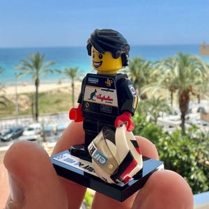 May include: A black racing driver minifigure with a yellow head and black hair, wearing a black suit with logos including Australian, Goodyear, and Olympus. The figure holds a white helmet with red and black accents on a black stand labeled Elio De Ang.