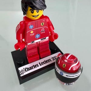 May include: A red racing driver minifigure with black hair, wearing a Ferrari team suit with HP, VGW, and Shell logos, standing on a black display base labeled Charles Leclerc 25, accompanied by a matching red and white racing helmet.