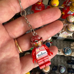 Könnte beinhalten: Ein roter und weißer Lego-Minifiguren-Schlüsselanhänger mit einem Marlboro-Logo auf der Vorderseite. Die Minifigur trägt einen roten Rennanzug und einen Helm mit einem Marlboro-Logo. Der Schlüsselanhänger ist an einer silbernen Kette befestigt.