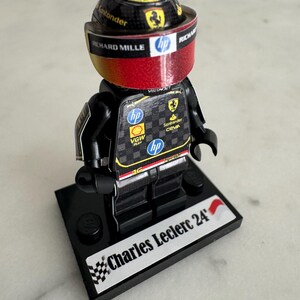 May include: A black Lego minifigure of a race car driver wearing a black and red racing suit and a black helmet with a Ferrari logo. The minifigure is standing on a black base with the words "Charles Leclerc 24'" printed on it.