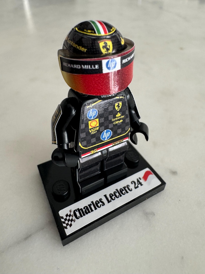 May include: A black Lego minifigure of a race car driver wearing a black and red racing suit and a black helmet with a Ferrari logo. The minifigure is standing on a black base with the words "Charles Leclerc 24'" printed on it.