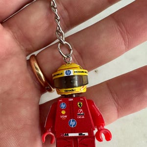 Può includere: Portachiavi Lego minifigure rosso con un casco giallo e loghi di sponsor come HP, Vistec, Richard Mille e Shell. La figura indossa una tuta da corsa rossa con il logo Ferrari.