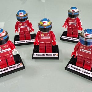 May include: Six Lego-style figurines of a Formula 1 driver, each on a black base. The figures wear red racing suits and helmets with blue, white, and yellow accents. Each base has the name "Fernando Alonso" and a number.