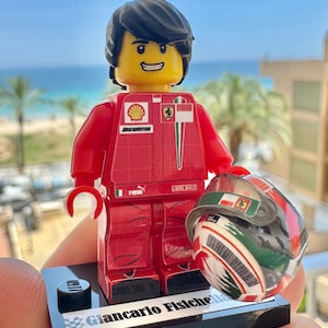 May include: A plastic minifigure of a race car driver wearing a red racing suit with sponsor logos including Shell, Bridgestone, and Puma. The figure has black hair, a yellow face, and holds a racing helmet. It stands on a black display base.