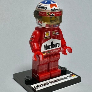 May include: A Lego figurine of Michael Schumacher in a red racing suit with Marlboro branding and a helmet featuring a star-studded design. The figurine stands on a black base with the name "Michael Schumacher 98" printed on it.