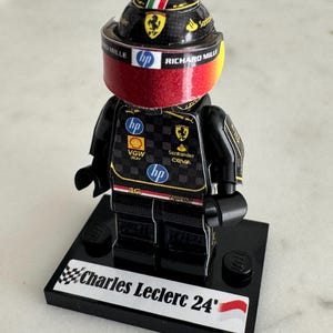 May include: A black Lego figurine of a race car driver wearing a black and red racing suit with a black helmet with a red visor. The figurine is standing on a black base with the words "Charles Leclerc 24'" printed on it.