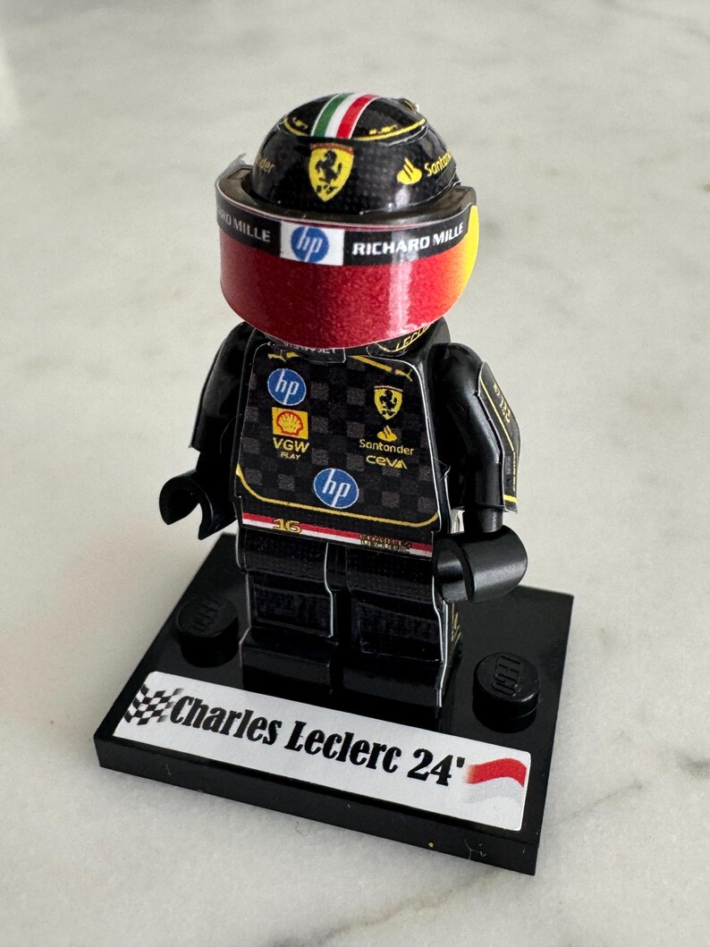 May include: A black Lego figurine of a race car driver wearing a black and red racing suit with a black helmet with a red visor. The figurine is standing on a black base with the words "Charles Leclerc 24'" printed on it.