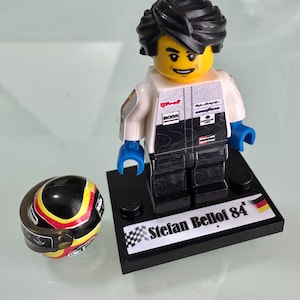 May include: A plastic minifigure of a race car driver with black hair, wearing a white and black racing suit with sponsor logos, standing on a black display base labeled Stefan Bellof 84 with a German flag, next to a black, red, and yellow helmet.