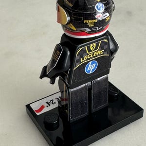 May include: A black and gold Lego minifigure of a race car driver wearing a black helmet with a red, white, and green Italian flag on the top. The figure is standing on a black base with a white and red Polish flag sticker. The figure's racing suit has the name "Leclerc" and the logo "HP" on the back.