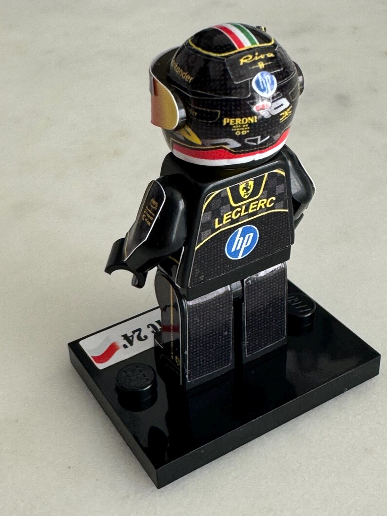 May include: A black and gold Lego minifigure of a race car driver wearing a black helmet with a red, white, and green Italian flag on the top. The figure is standing on a black base with a white and red Polish flag sticker. The figure's racing suit has the name "Leclerc" and the logo "HP" on the back.