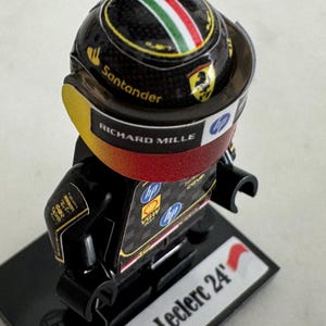 May include: A black Lego minifigure of a race car driver wearing a black and yellow helmet with the words "Richard Mille" and "Santander" on it. The figure is standing on a black base with the words "Leclerc 24" printed on it.