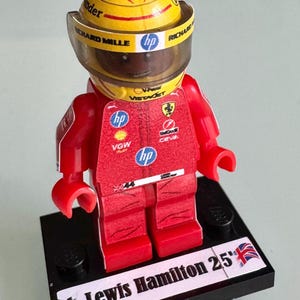 May include: A red Lego figurine of a race car driver wearing a yellow helmet with the words "Richard Mille" and "hp" on it. The figurine is standing on a black base with the words "Lewis Hamilton 25'" and a Union Jack flag.