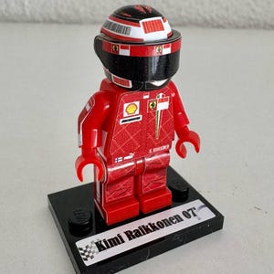 May include: A red, white, and black Lego figurine of a race car driver wearing a helmet and a racing suit with the name "Kimi Raikkonen 07" on the base.