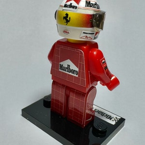 May include: A red and white Lego figure of a race car driver, wearing a helmet with the Marlboro logo and a visor. The figure is on a black base with the name "Michael" printed on it.
