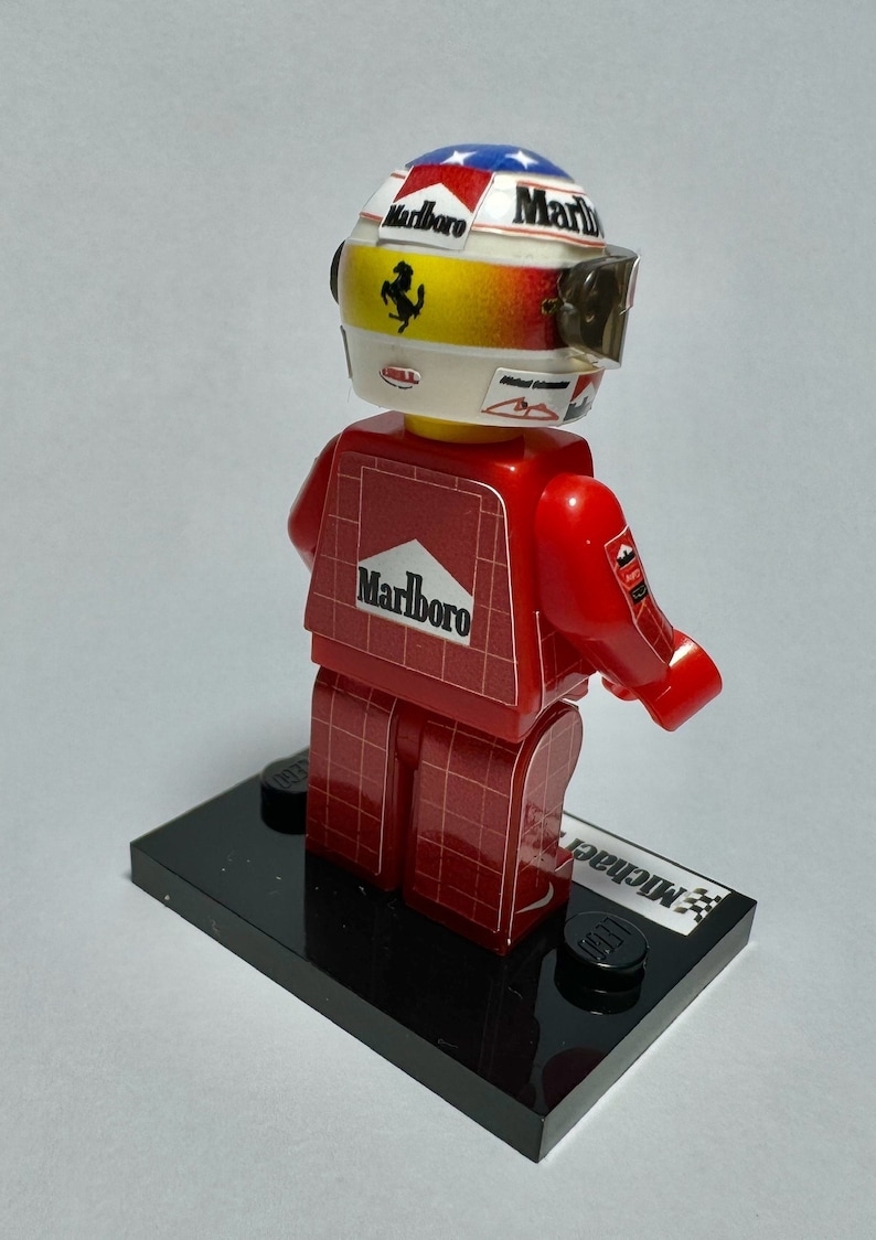 May include: A red and white Lego figure of a race car driver, wearing a helmet with the Marlboro logo and a visor. The figure is on a black base with the name "Michael" printed on it.