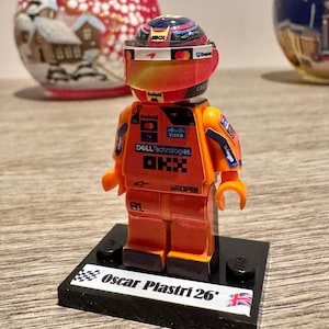 Op de afbeelding: Een Lego-figuur van een Formule 1-coureur in een oranje racepak, helm en vizier. De figuur staat op een zwart voetstuk met de naam "Oscar Piastri 26" en een Britse vlag. De achtergrond bevat twee decoratieve glazen bollen.