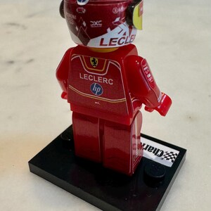 May include: A red and white Lego minifigure of a race car driver wearing a helmet with the name "Leclerc" on it. The figure is standing on a black base with a checkered flag.