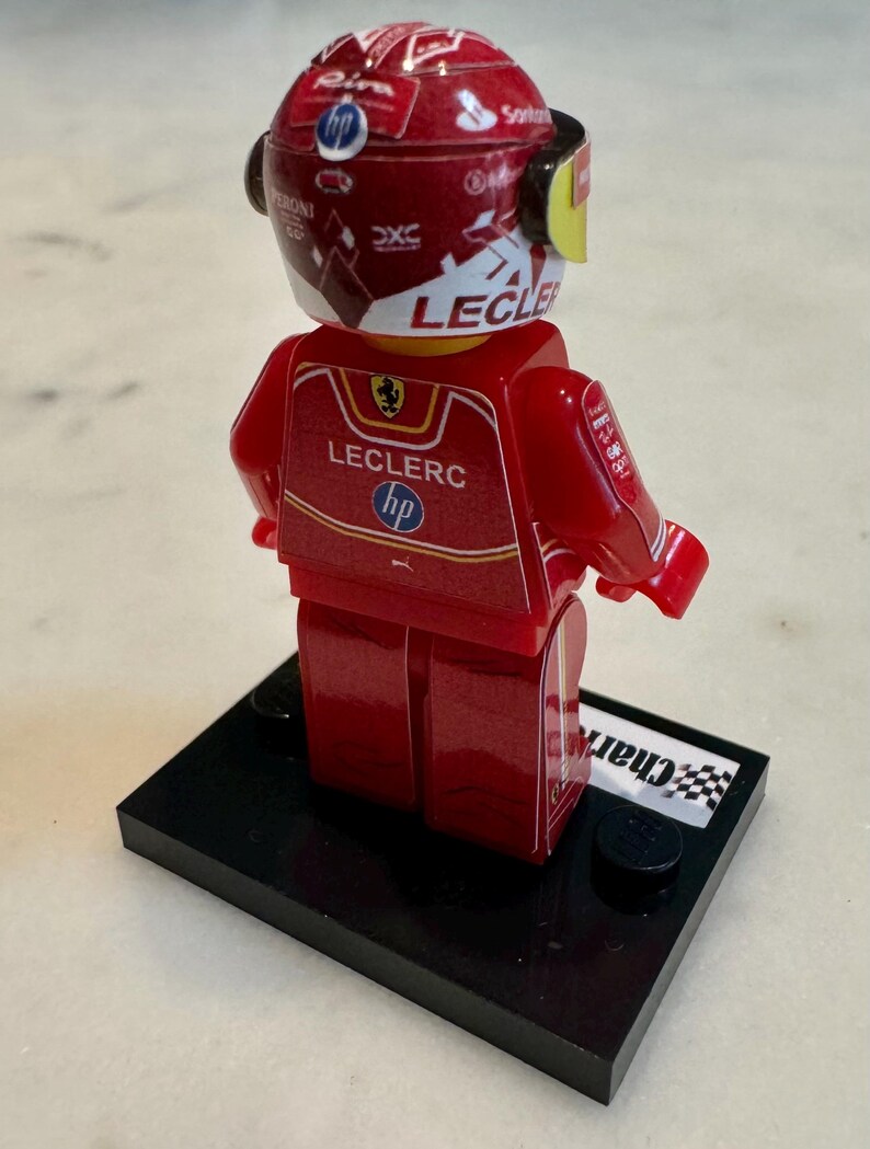 May include: A red and white Lego minifigure of a race car driver wearing a helmet with the name "Leclerc" on it. The figure is standing on a black base with a checkered flag.