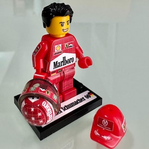 May include: A plastic toy figure of a race car driver wearing a red racing suit with Marlboro branding, standing on a black display base labeled Michael Schumacher 00. The figure holds a red helmet, and a separate red cap is placed next to the base.