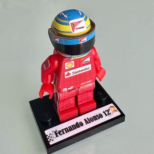 Può includere: Una statuetta Lego di un pilota di Formula 1, con tuta da corsa rossa e casco con dettagli blu, gialli e bianchi. La statuetta è su una base nera con il nome "Fernando Alonso 12" stampato.