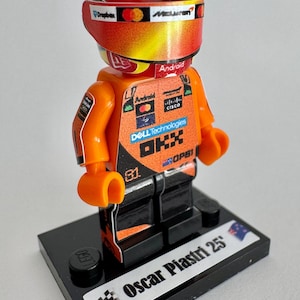 Op de afbeelding: Een Lego-figuur van een raceautocoureur die een oranje en zwart racepak draagt met sponsorlogo's, waaronder "DHL Technologies", "Android" en "McLaren". Het figuur staat op een zwarte basis met de tekst "Oscar Piastri 25" en een zwart-wit geblokt ontwerp.