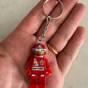Può includere: Figurino Lego rosso di un pilota di auto da corsa che indossa un casco con il logo Marlboro e il testo "Niki Lauda" su di esso. Il figurino tiene una chiave inglese e ha la parola "parmalat" stampata sulla parte anteriore della sua tuta da corsa.