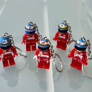 Puede incluir: Cinco llaveros en miniatura de pilotos de carreras de color rojo y azul con llaveros plateados. Cada llavero presenta un piloto con un traje de carreras rojo y un casco azul y blanco.