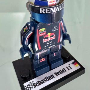 Peut inclure: Figurine Lego de Sebastian Vettel dans une voiture de Formule 1 Red Bull Racing. La figurine porte un casque bleu avec le logo Renault et le logo Red Bull sur la poitrine. La base est noire et porte le nom "Sebastian Vettel 11".