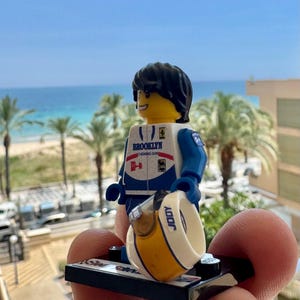 May include: A plastic minifigure with black hair and a yellow face, wearing a white and blue racing suit with the text BROOKLYN CHEWING GUM printed on the chest. The figure holds a white helmet with a yellow stripe and the text JODY on the side.