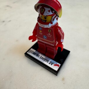 May include: A red and white Lego minifigure of a race car driver wearing a helmet with the words "Richard Mille" on it. The figure is standing on a black base with the words "Charles Leclerc 24" printed on it.