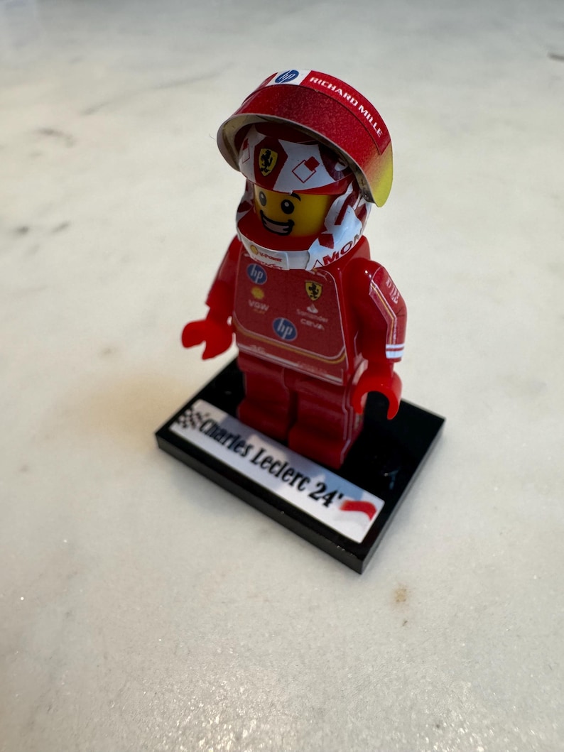 May include: A red and white Lego minifigure of a race car driver wearing a helmet with the words "Richard Mille" on it. The figure is standing on a black base with the words "Charles Leclerc 24" printed on it.