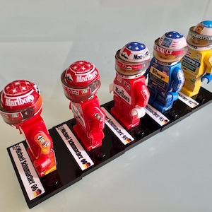Könnte beinhalten: Fünf Miniatur-Lego-Figuren von Rennfahrern, jede Figur trägt einen roten Rennanzug und einen Helm mit einem Marlboro-Logo. Die Figuren werden auf einem schwarzen Ständer mit weißen Textetiketten präsentiert, die "Michael Schumacher 98", "Michael Schumacher 00", "Michael Schumacher 06", "humacher 95" und "macher 92" lauten.