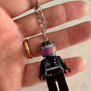May include: Black Lego minifigure keychain with a green and white racing suit. The figure has a helmet with a Petronas logo and a purple and pink iridescent visor. The figure also has a Petronas logo on the chest and a UBS logo on the arm.