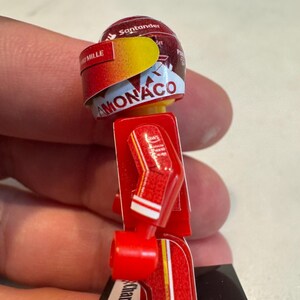 May include: A red and yellow Lego minifigure of a race car driver wearing a helmet with the words "Santander" and "Monaco" on it. The figure is wearing a red racing suit with white accents and the name "Charles Leclerc" on the leg.