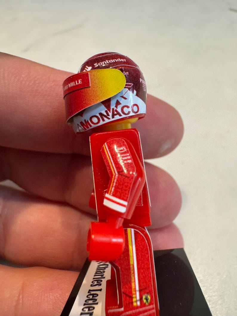 May include: A red and yellow Lego minifigure of a race car driver wearing a helmet with the words "Santander" and "Monaco" on it. The figure is wearing a red racing suit with white accents and the name "Charles Leclerc" on the leg.