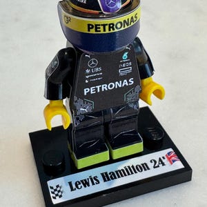 May include: A black Lego figurine of a race car driver wearing a black and green racing suit with the word "PETRONAS" on the chest. The figurine is wearing a helmet with a purple and yellow design and the word "PETRONAS" on the front. The figurine is standing on a black base with the words "Lewis Hamilton 24" and a Union Jack flag.