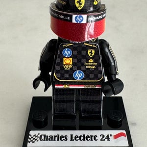 May include: A black Lego figurine of a race car driver wearing a red and black racing suit with a black helmet and visor. The figurine is standing on a black base with a white label that reads "Charles Leclerc 24".