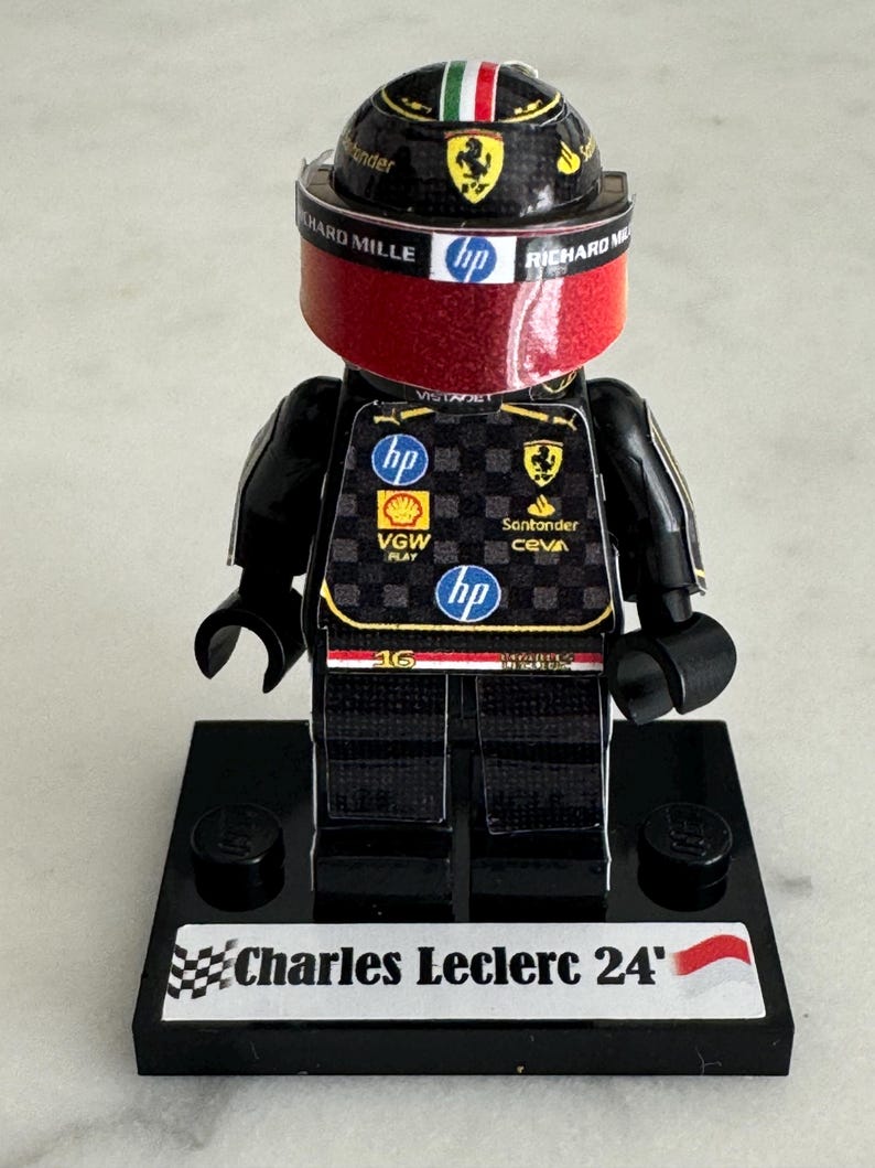 May include: A black Lego figurine of a race car driver wearing a red and black racing suit with a black helmet and visor. The figurine is standing on a black base with a white label that reads "Charles Leclerc 24".
