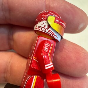 May include: A red and white Lego minifigure of a race car driver wearing a red helmet with the name "Leclerc" and the sponsor "Santander" on it.