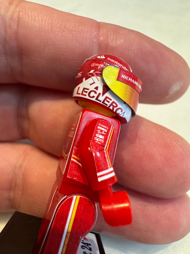 May include: A red and white Lego minifigure of a race car driver wearing a red helmet with the name "Leclerc" and the sponsor "Santander" on it.