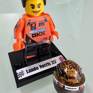 May include: A Lando Norris minifigure wearing an orange and black racing suit with sponsor logos including Android, Cisco, Dell Technologies, and OKX. The figure stands on a black display base labeled Lando Norris 25 with a matching racing helmet.