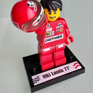 May include: A plastic minifigure of a race car driver wearing a red racing suit with various sponsor logos, including Parmalat and Marlboro. The figure holds a red racing helmet and stands on a black rectangular base labeled Niki Lauda 77.