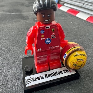 May include: A LEGO minifigure of a race car driver wearing a red racing suit with various sponsor logos, the number 44, and the name Lewis. The figure holds a yellow racing helmet and stands on a black display base labeled Lewis Hamilton 26.