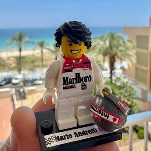 May include: A Lego minifigure of a race car driver with black hair and a yellow head, wearing a white racing suit with red accents and Marlboro branding. The figure stands on a black display base labeled Mario Andretti and holds a grey helmet.