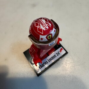 May include: A red and white Lego minifigure of a race car driver wearing a red helmet with a white Ferrari logo and the text "Scuderia Ferrari" on it. The minifigure is standing on a black base with the text "Charles Leclerc 24" on it.